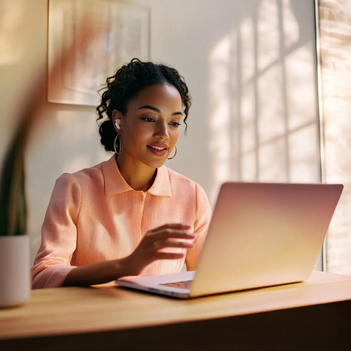 A professional woman with curly hair wearing a peach blouse and wireless earbuds, smiling while engaging in a RingCentral video meeting on her laptop in a bright, modern home office.