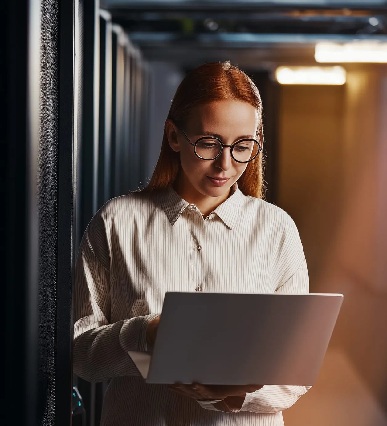 A woman stands inside an office, typing with one hand and holding the laptop with another.