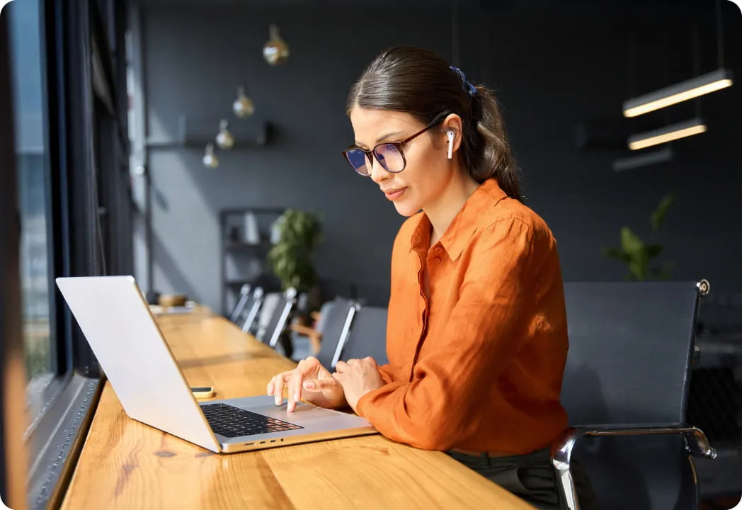 A woman wearing glasses sits at a table, focused on her laptop while working or studying.